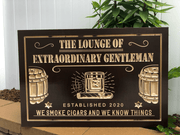 A rectangular dark brown wooden plaque with raised gold lettering and illustrations of cigar barrels, displayed outdoors against a white paneled wall with green plants above.